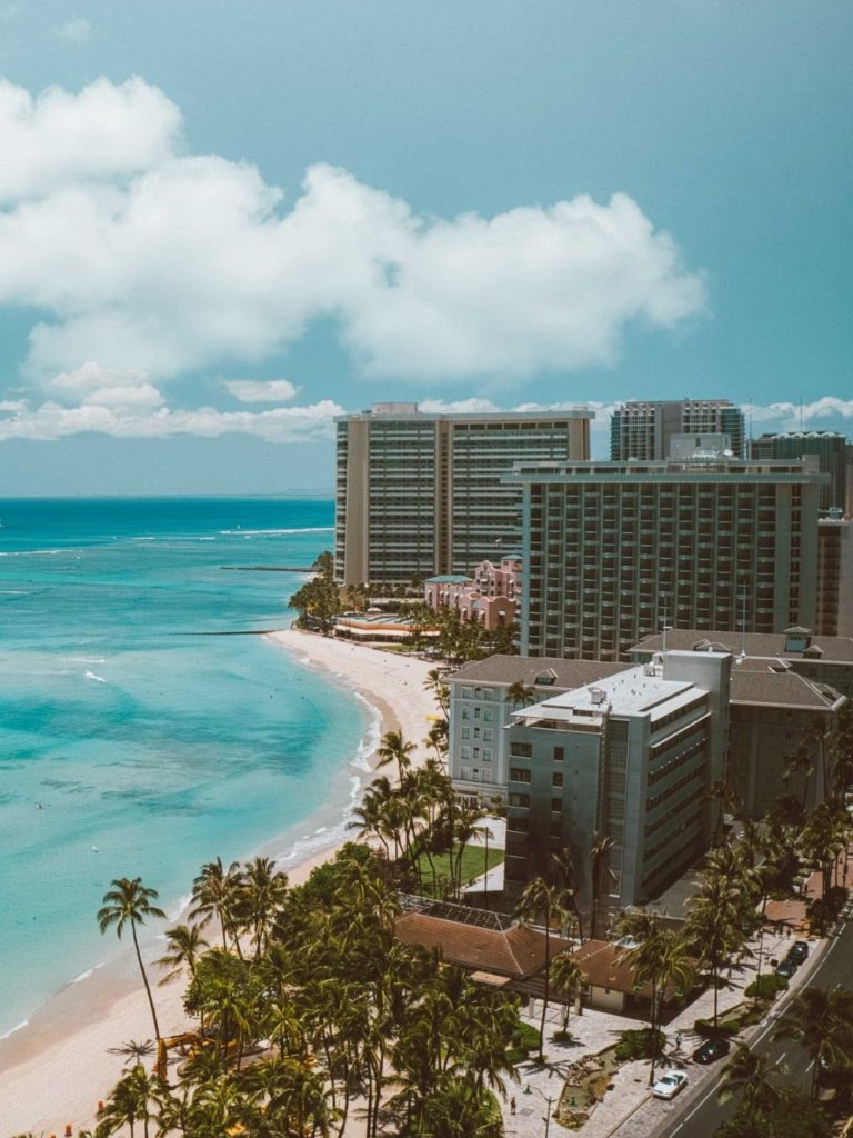 Travelowpay Vacation Coastal view featuring buildings along a sandy beach and turquoise water under a blue sky.